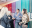 Three women are seen in a social setting, with the central figure wearing a prominent light blue faux fur jacket and patterned jeans, while the other two women wear dark and leopard-print outerwear. They are standing in front of a colorful striped wall with red, purple, and teal panels.