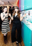 Two women posing in a hallway with colorful walls. The woman on the left wears a black, white, and gray striped knit dress with brown knee-high boots.